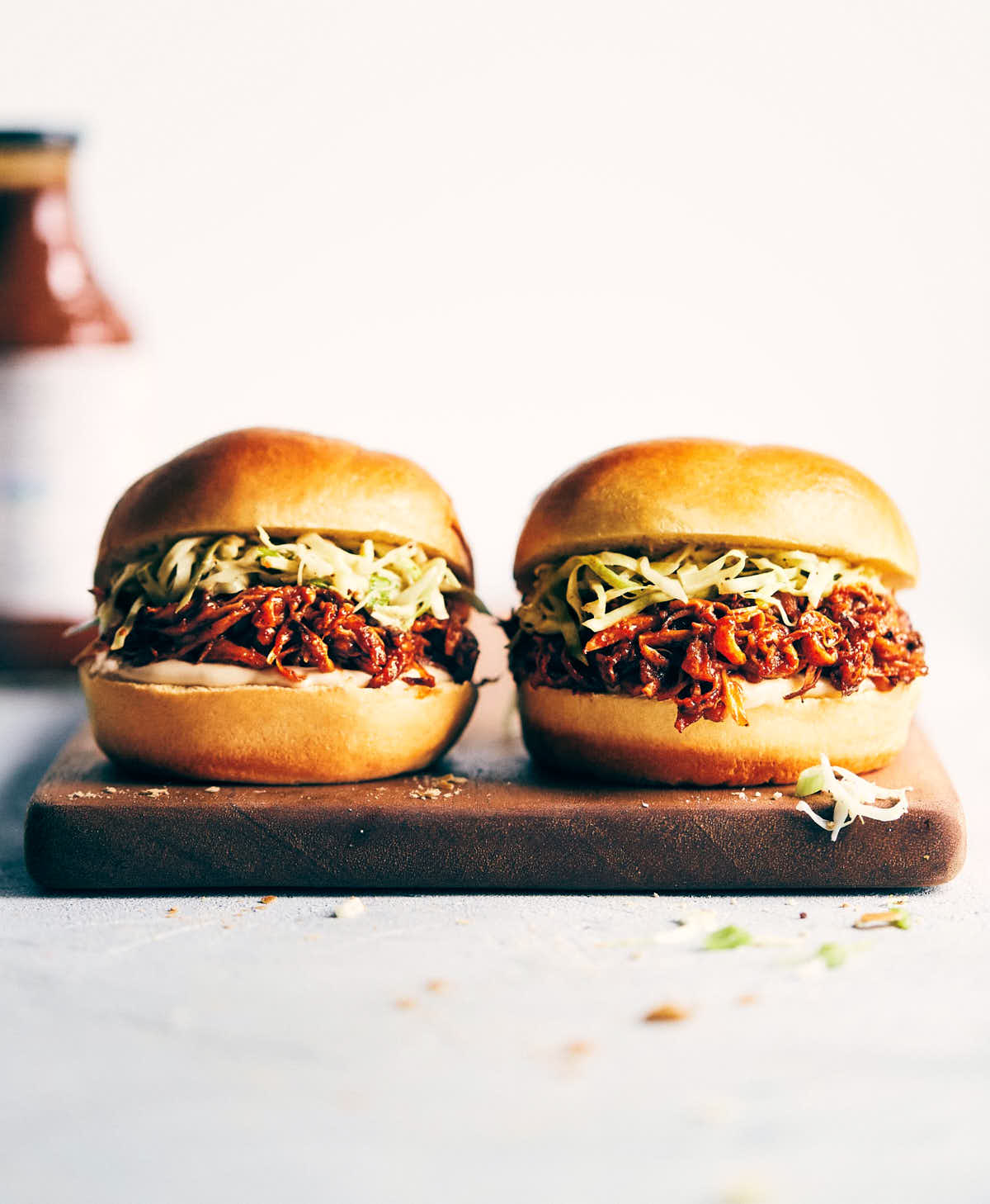 Two barbecue pulled mushroom sandwiches on a serving board with barbecue sauce in the background