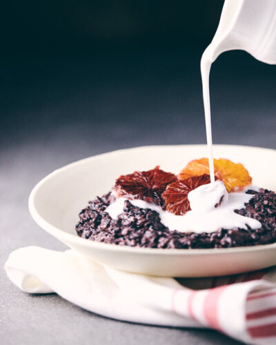 Pouring coconut milk over black rice pudding with blood oranges