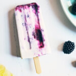 Blackberry cheesecake popsicle sitting on a counter with blackberries and cashews surrounding