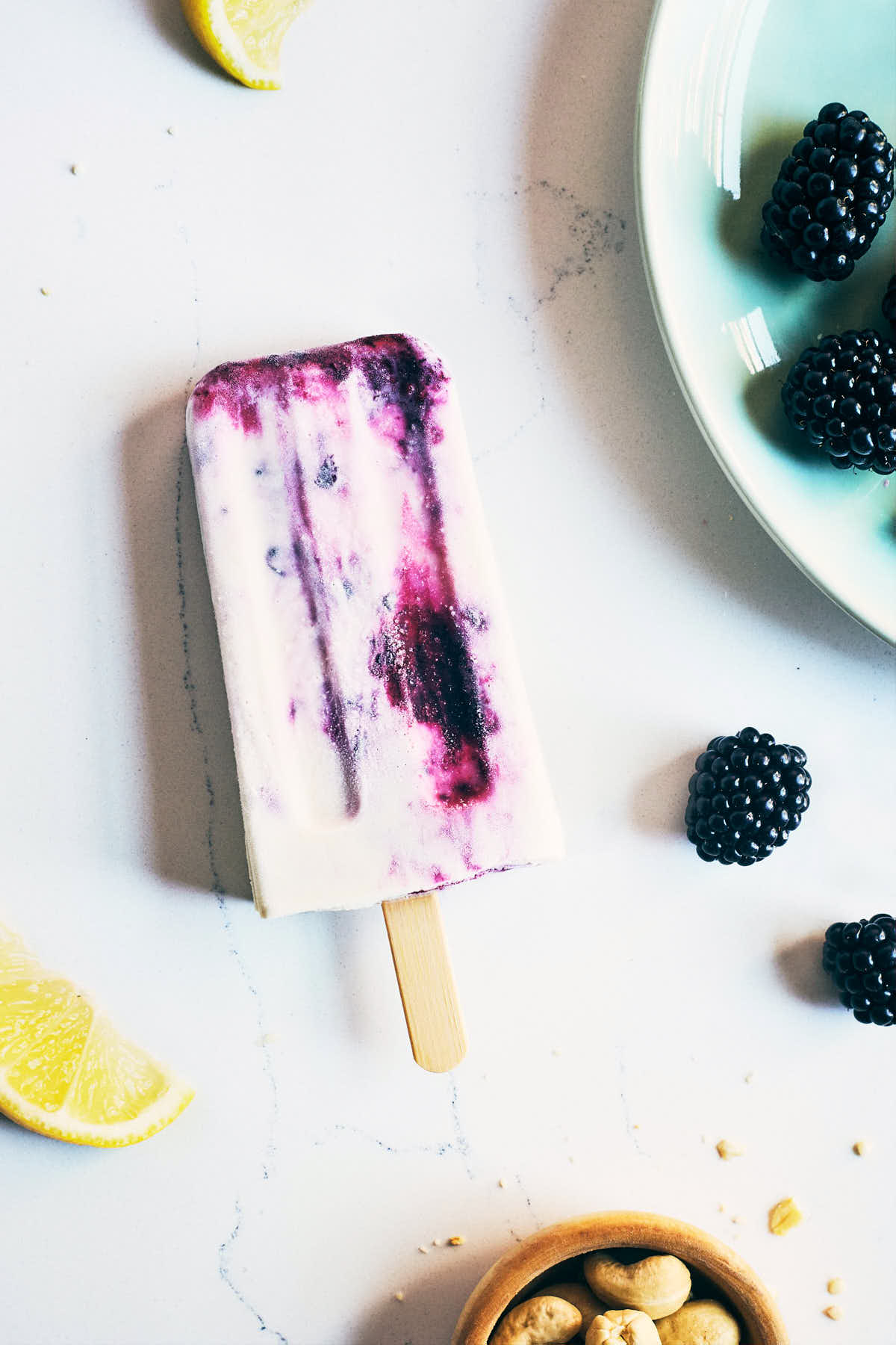 Finished blackberry cheesecake popsicle sitting on a counter with blackberries, lemons and cashews surrounding