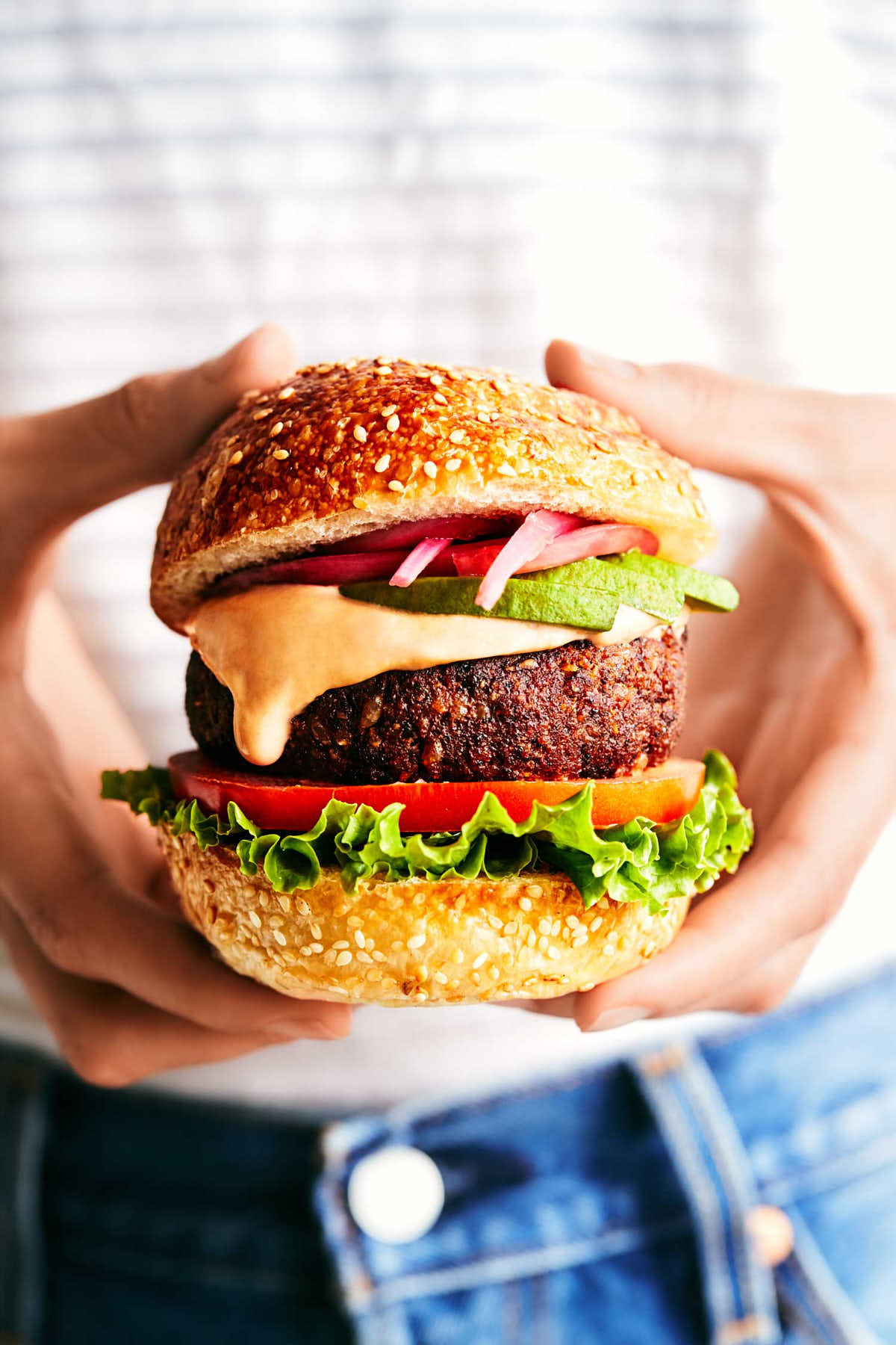 Chipotle black bean burger with avocado, pickled red onions and chipotle mayo being held in front of a white shirt and denim jeans