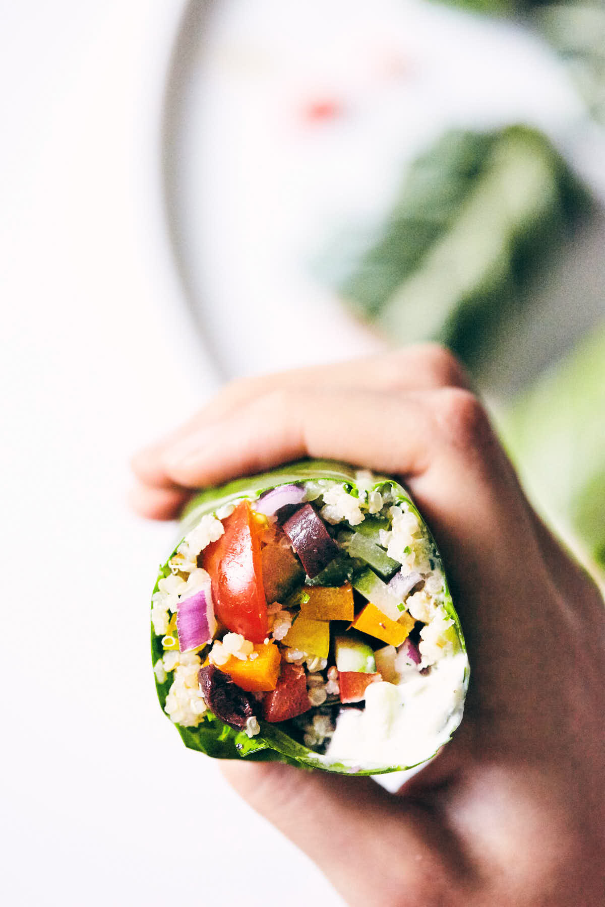 Hand holding green collard wrap sliced in half with peppers, tzatziki, cucumber, red onion and olives visible inside