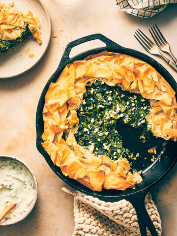Cast iron pan on the counter with spanakopita slice cut out and served with tzatziki