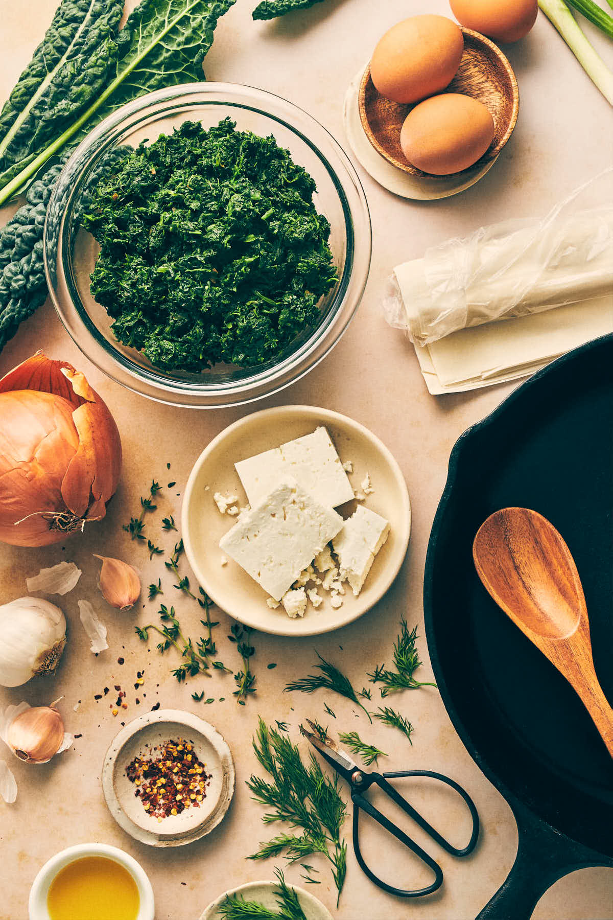 Spanakopita ingredients like spinach, feta, dill, garlic and eggs laid out on the counter with a cast iron pan