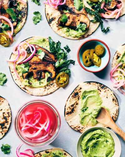 Wide shot of five smoky mushroom tacos on a counter being dressed with toppings like smoky mushrooms, avocado crema, pickled red onions and freshly chopped kale