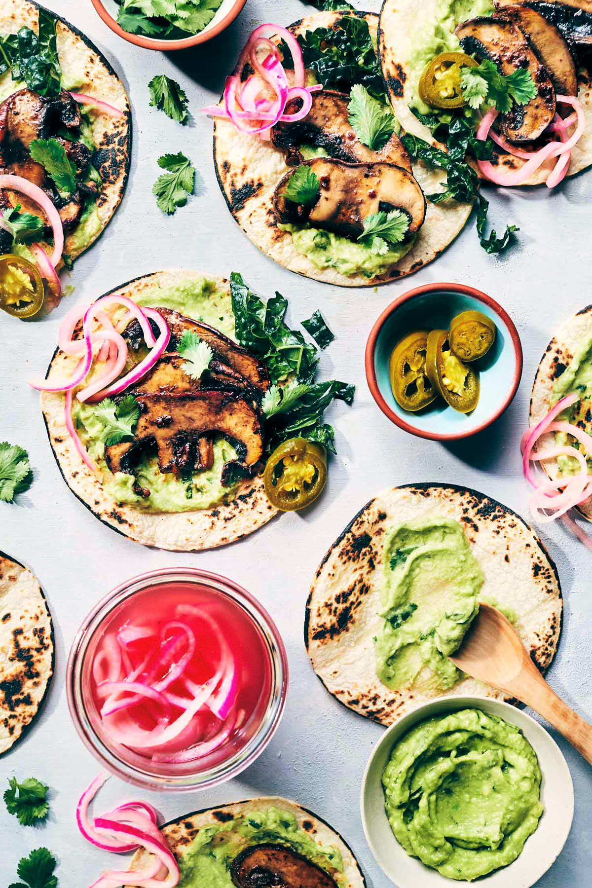 Wide shot of five smoky mushroom tacos on a counter being dressed with toppings like smoky mushrooms, avocado crema, pickled red onions and freshly chopped kale