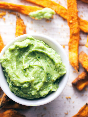 Spiced sweet potato fries spread across white wooden background with avocado dip