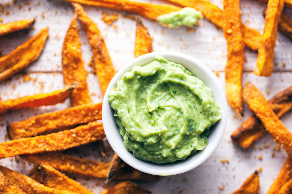 Spiced sweet potato fries spread across white wooden background with avocado dip