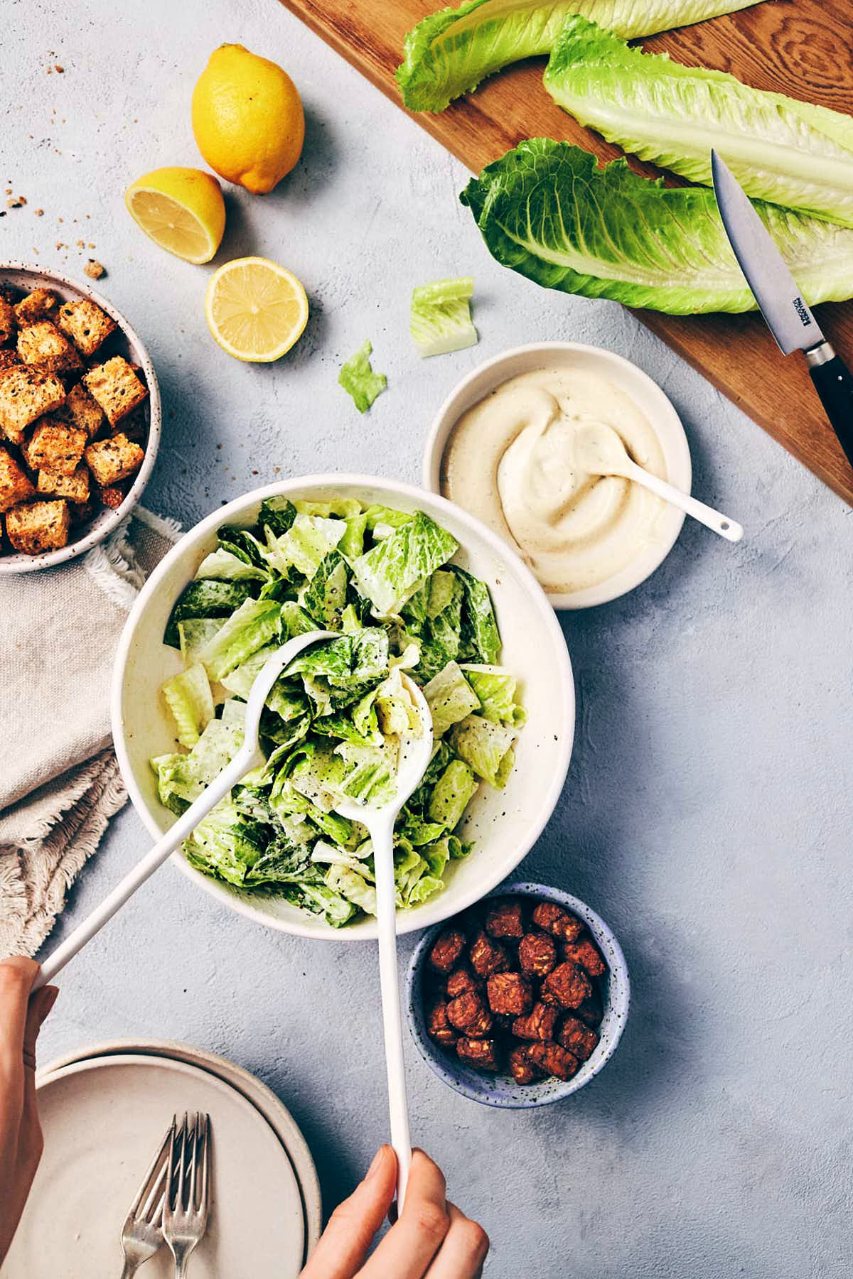 Hands tossing Caesar lettuce with dressing before being topped with tempeh and crispy croutons