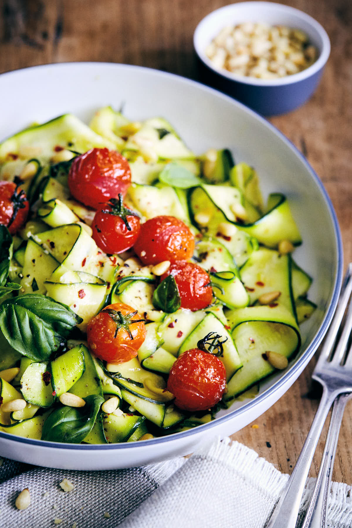 Zucchini pappardelle with roasted tomatoes and basil in a bowl with pine nuts on the side