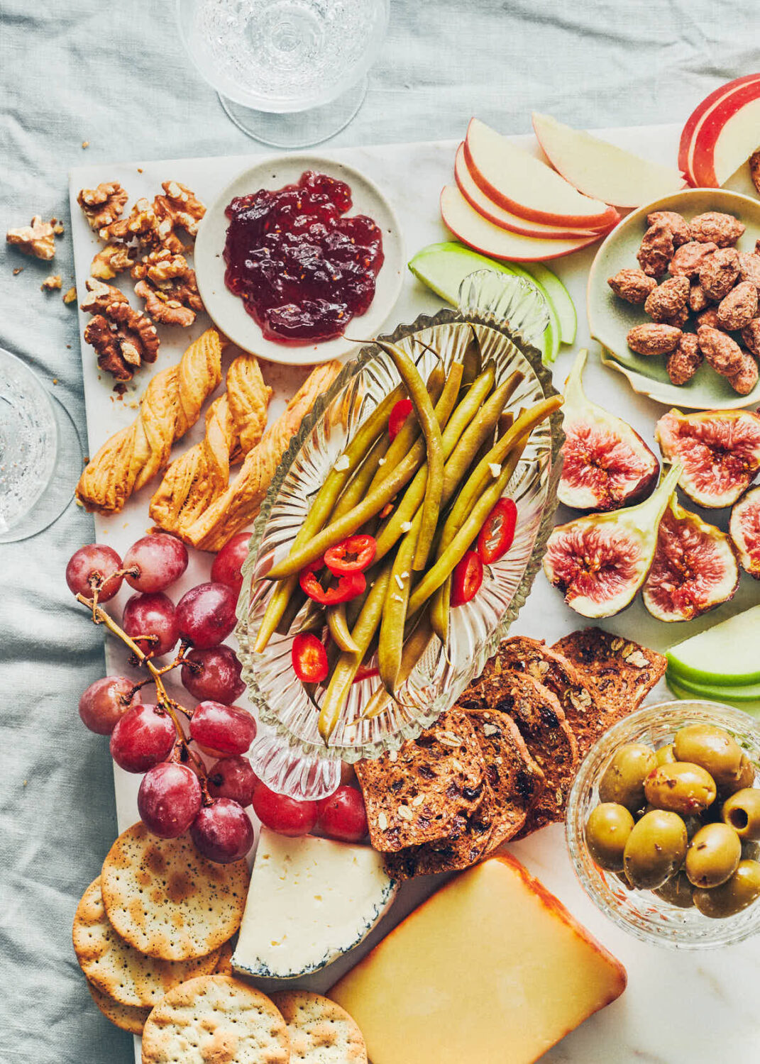 Snack board with fruit, crackers, olives, jam and pickled green beans on a table