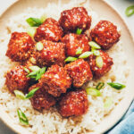 Crispy thai sweet chili tofu sitting in a bowl of white rice with scallions/green onions sprinkled on top