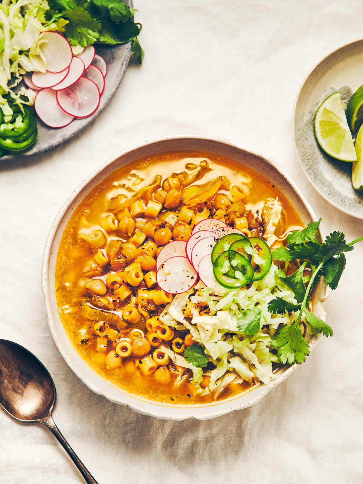 A bowl of chickpea (garbanzo bean) noodle soup topped with shredded lettuce, jalapenos and radish, sitting on a white linen tablecloth