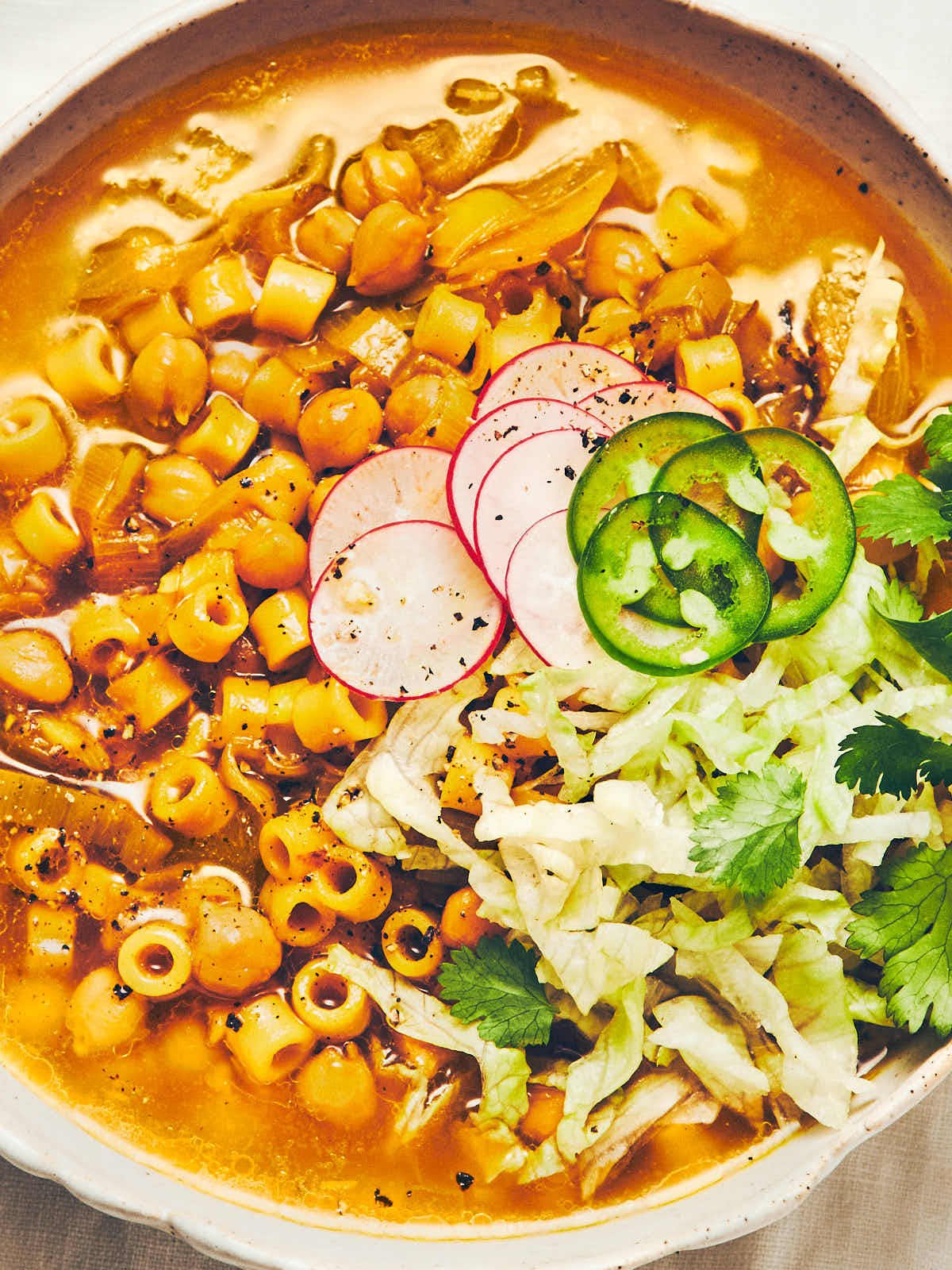 An overhead photo of a bowl filled with golden chickpea noodle soup, topped with shredded iceberg lettuce, radishes, and jalapeno peppers