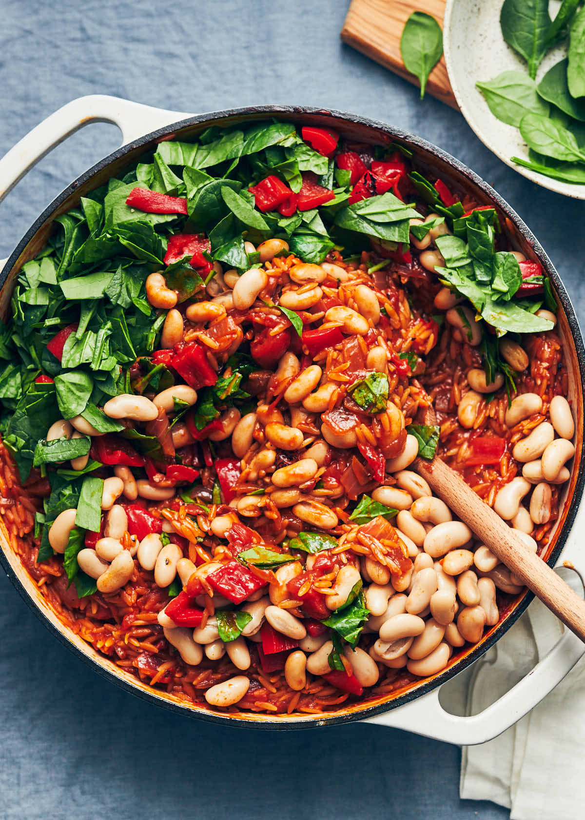 A wooden stirring spoon mixing white cannellini beans into a pot of tomato harissa orzo with spinach
