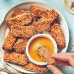 A plate of crispy air fryer tofu sticks, with a stick being dipped into a bowl of honey mustard sauce.