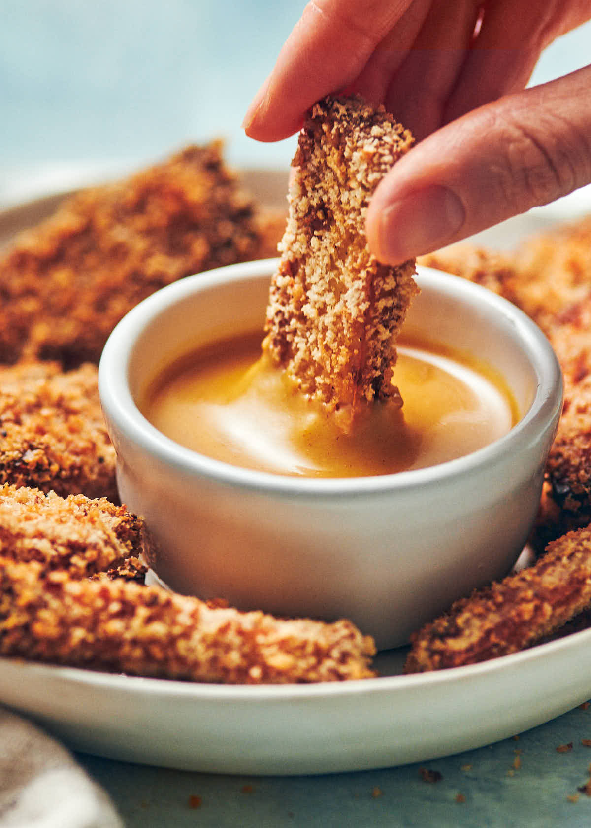 A hand holding a piece of crispy panko crusted tofu, dipping it into a bowl of honey mustard sauce.