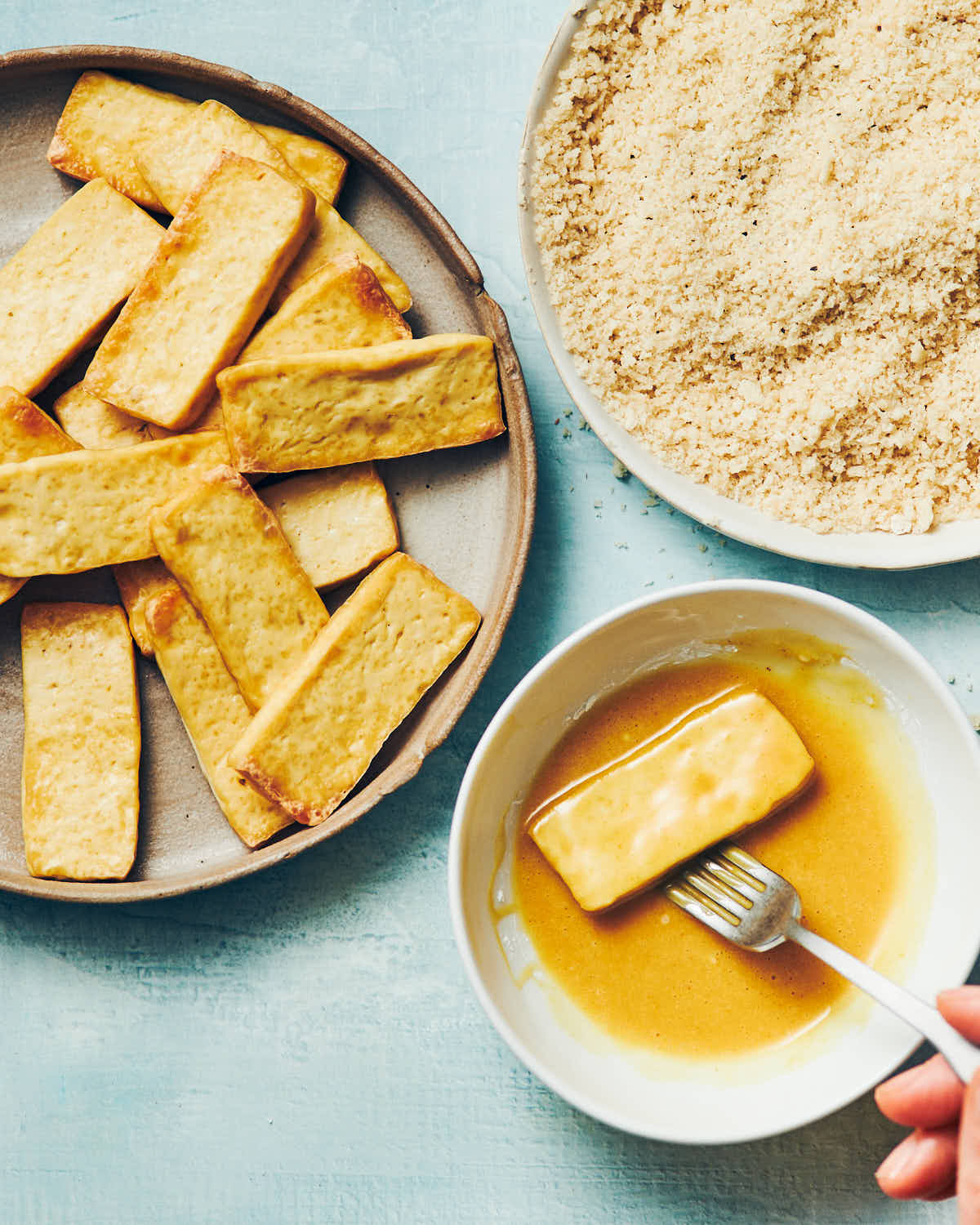 A fork dipping a slice of tofu in homemade honey mustard sauce, with a plate of panko breadcrumbs nearby