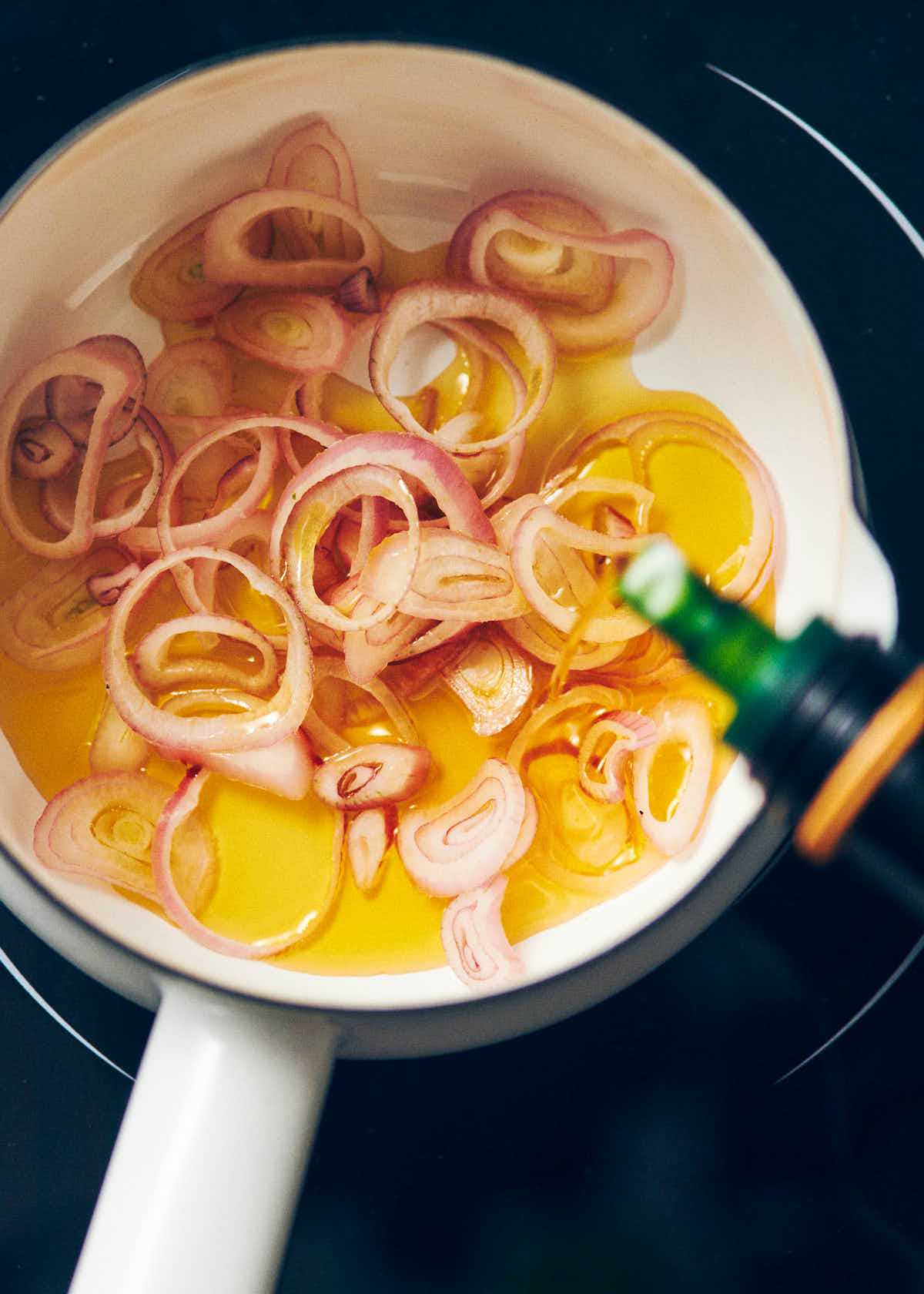 Olive oil being poured over thinly sliced shallots in a white enamel saucepan.