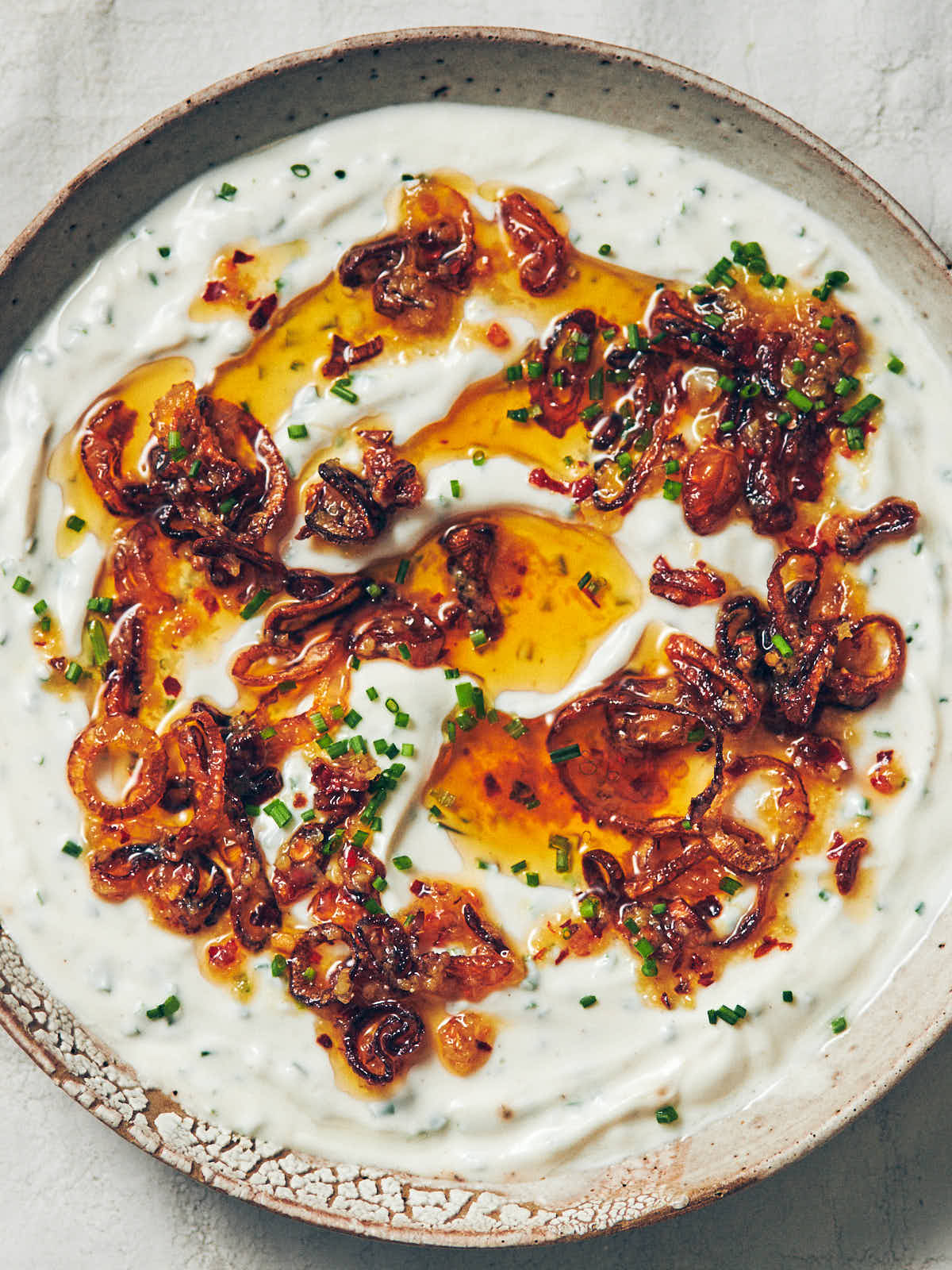 A ceramic serving bowl of Greek yogurt onion dip topped with chili garlic oil, fresh chives, and fried shallots