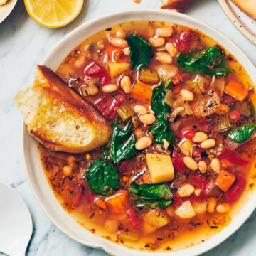 A bowl of Instant Pot Navy Bean Soup with a piece of bread sitting on the edge of the bowl, with a white spoon and sliced lemon