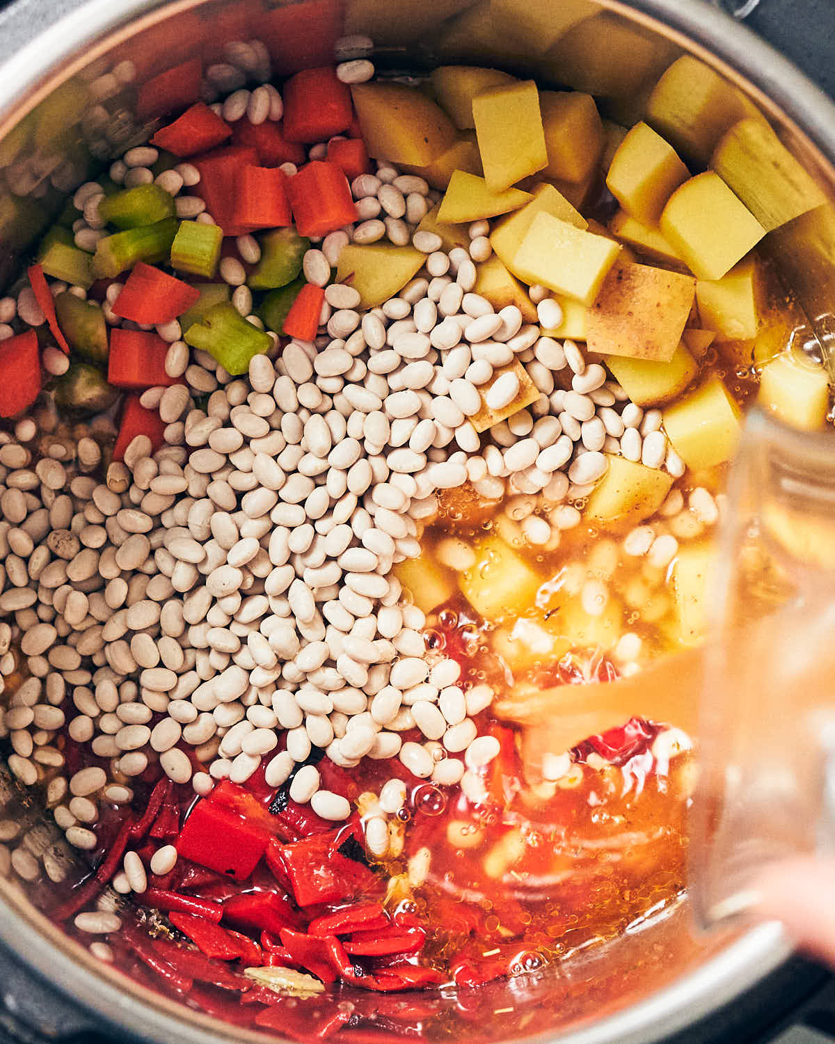 An Instant Pot with navy beans, roasted red peppers, carrots, celery, potatoes, and vegetable stock being poured in
