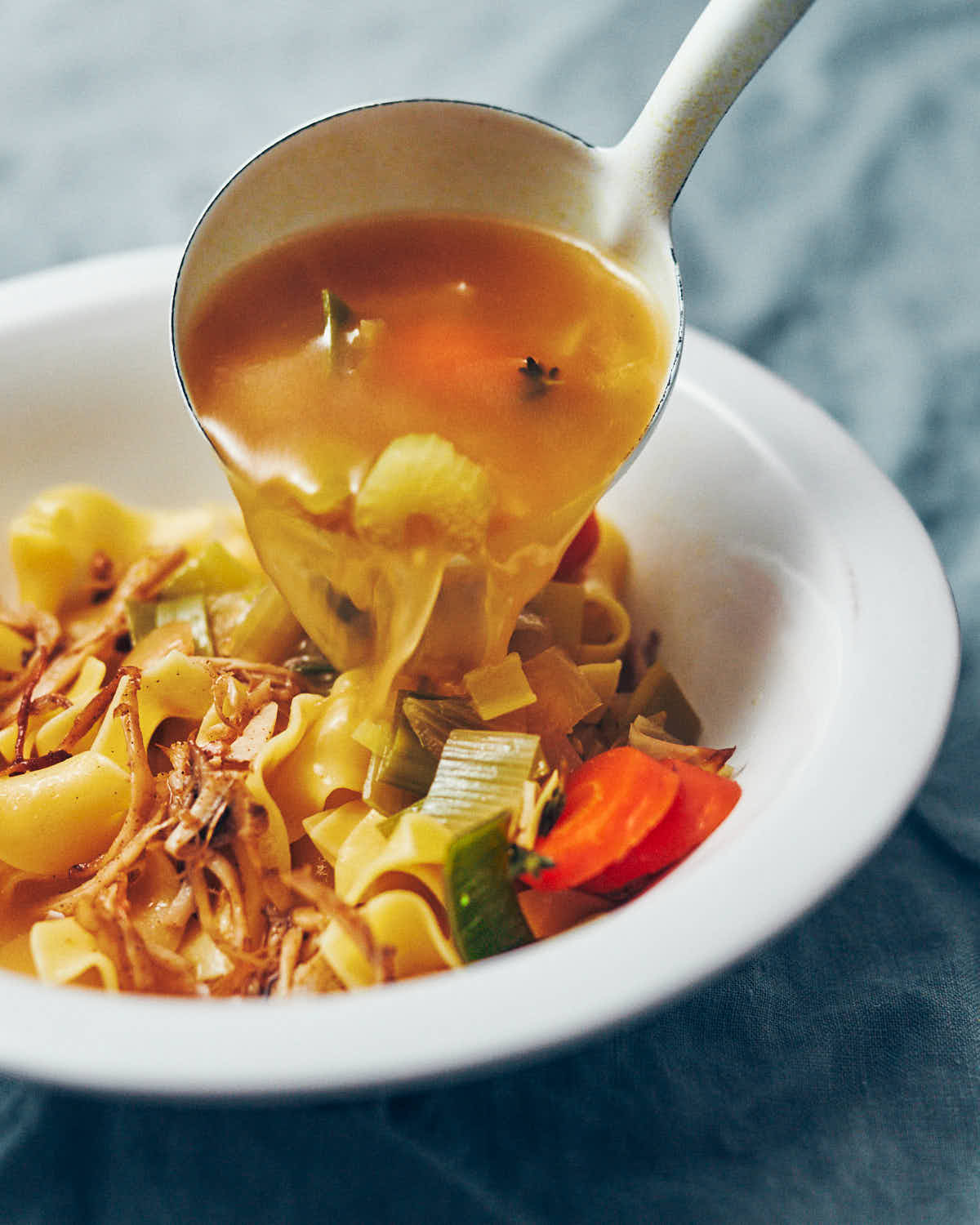 A white enamel ladle spooning vegan chicken noodle soup into a white serving bowl.