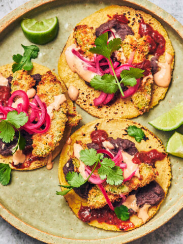 Three vegan air fried avocado tacos on a plate, topped with pickled onion.
