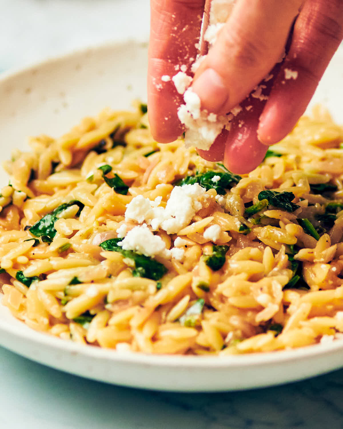 A hand sprinkling feta cheese over a plate of One Pan Lemon Spinach Orzo.