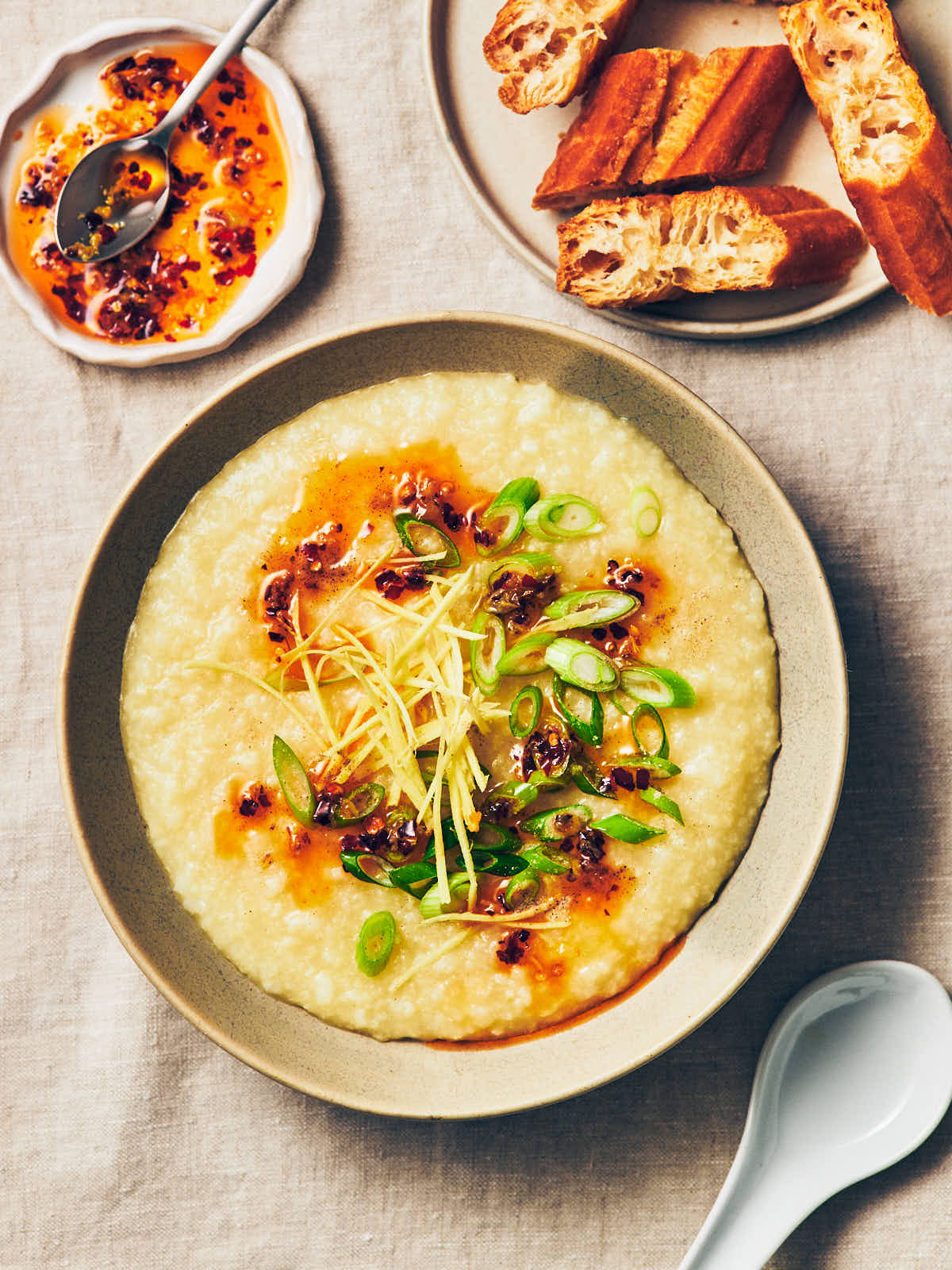 A bowl of vegan congee (rice porridge) topped with chili garlic oil, ginger, and scallions sitting on a table, next to a small plate of Chinese doughnut (Youtiao).