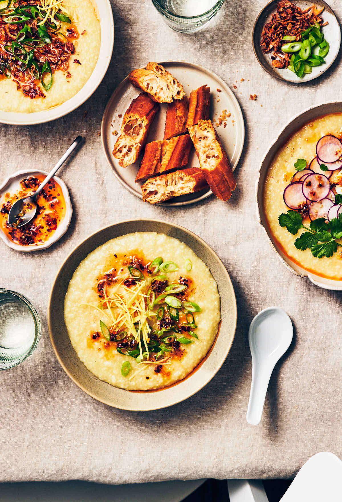 3 bowls of vegan congee with different toppings on a dining table, with a plate of Chinese donuts (youtiao) and glasses of water