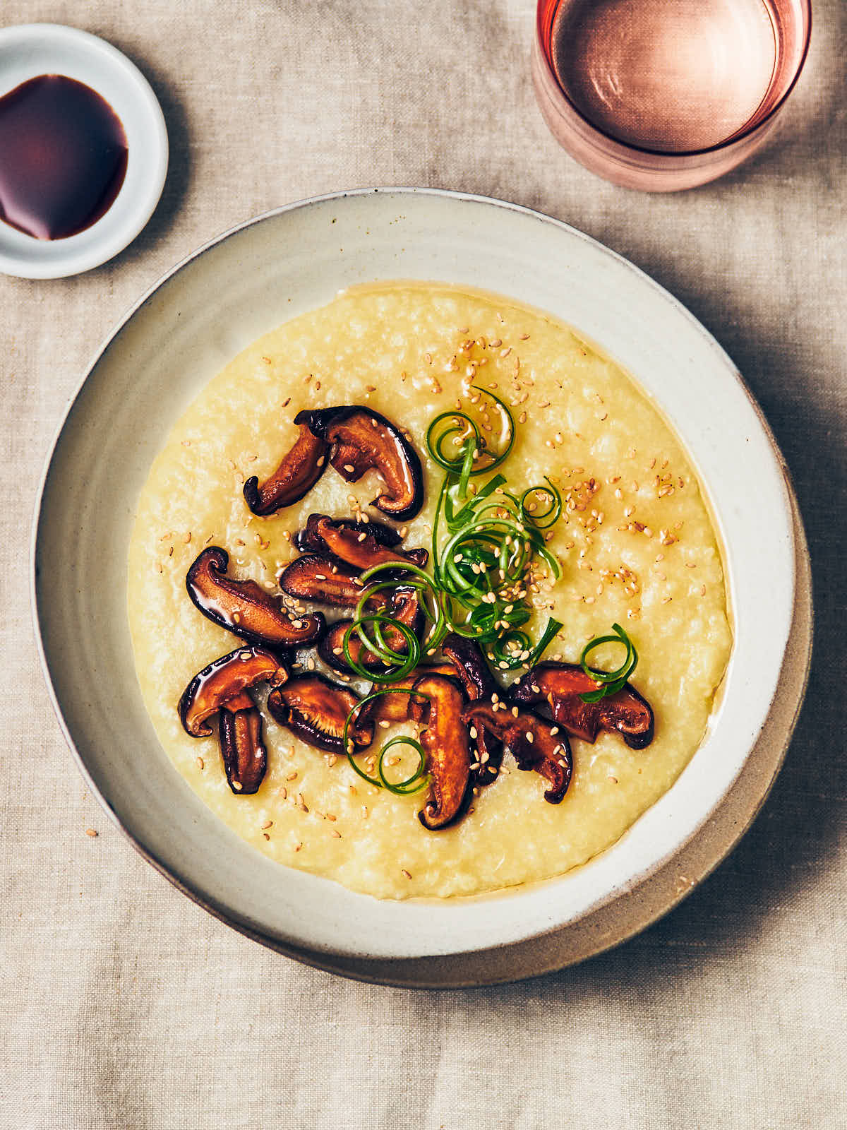 A bowl of congee with sauteed mushrooms, toasted sesame oil, sesame seeds, and scallions on top with a small dish of soy sauce on the side.