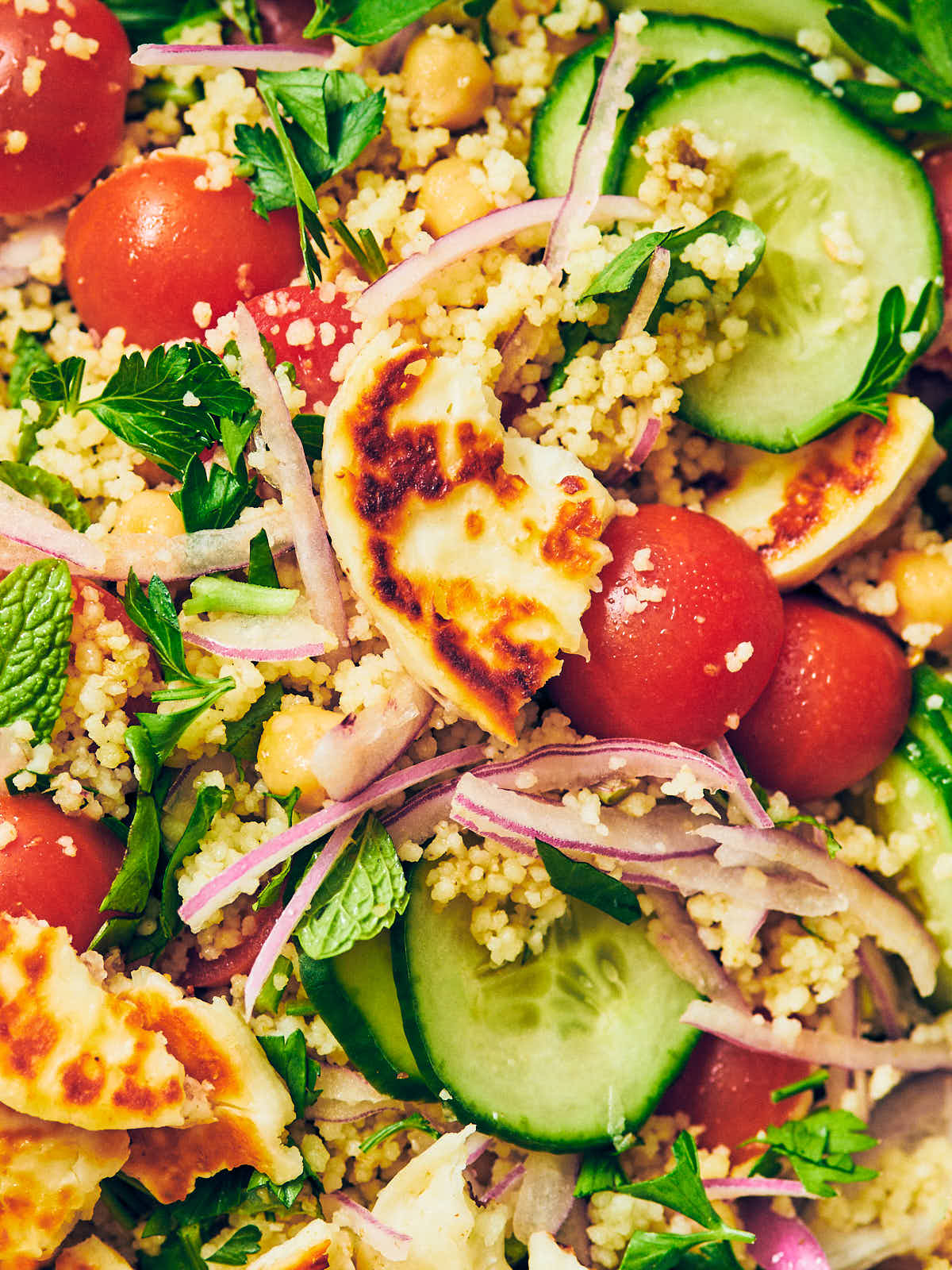 Halloumi Couscous Salad with sliced cucumbers, red onion, tomatoes, parsley, basil, mint, and seared halloumi cheese.