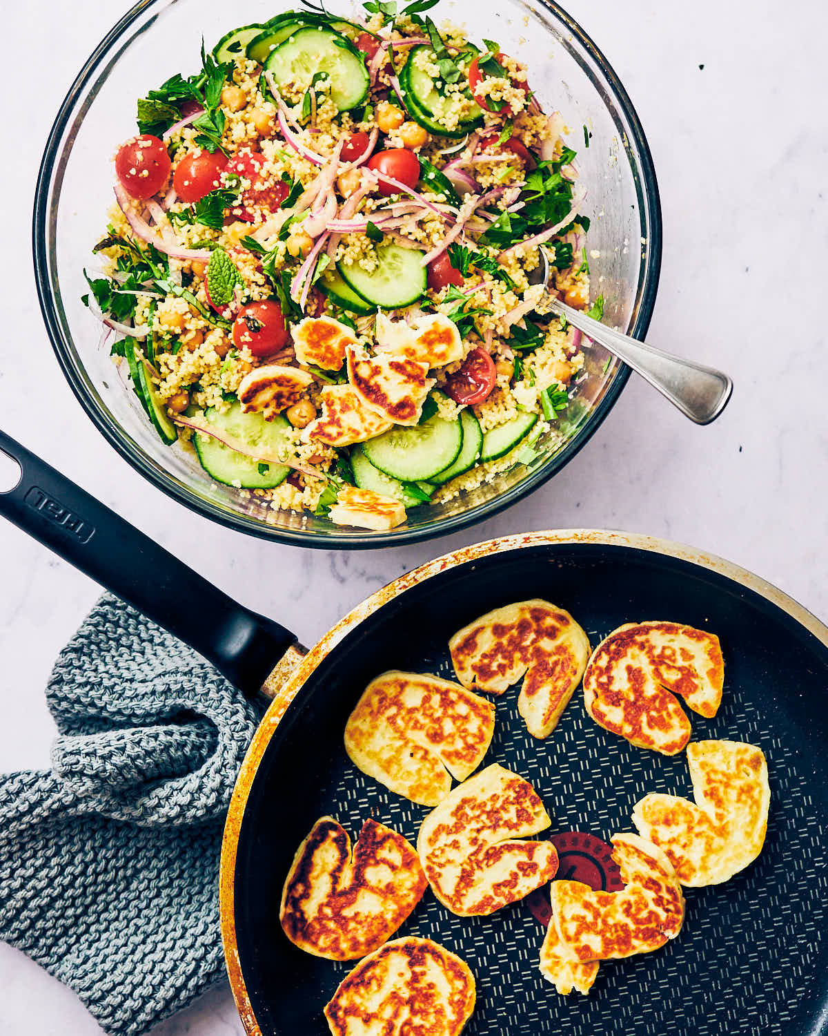 A skillet of seared halloumi, being added to a serving bowl of Halloumi Couscous Salad with Lemon.