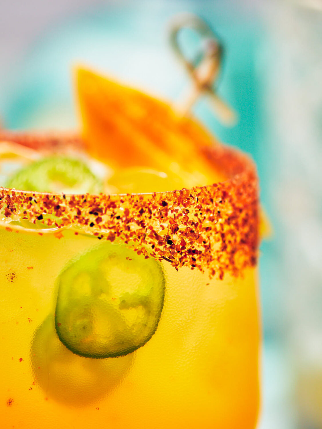 Closeup of a tajin rimmed glass filled with mango margarita on the rocks with jalapenos. 