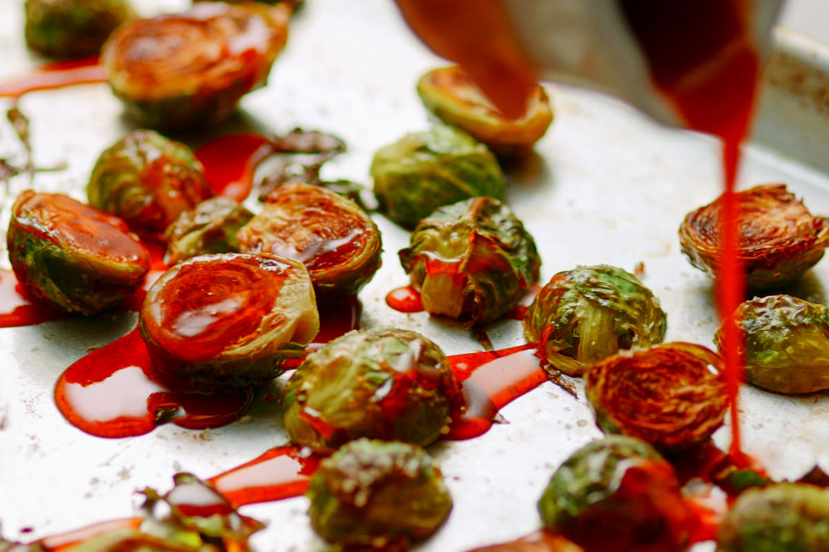 Drizzling maple sriracha sauce over roasted brussels sprouts on baking sheet.