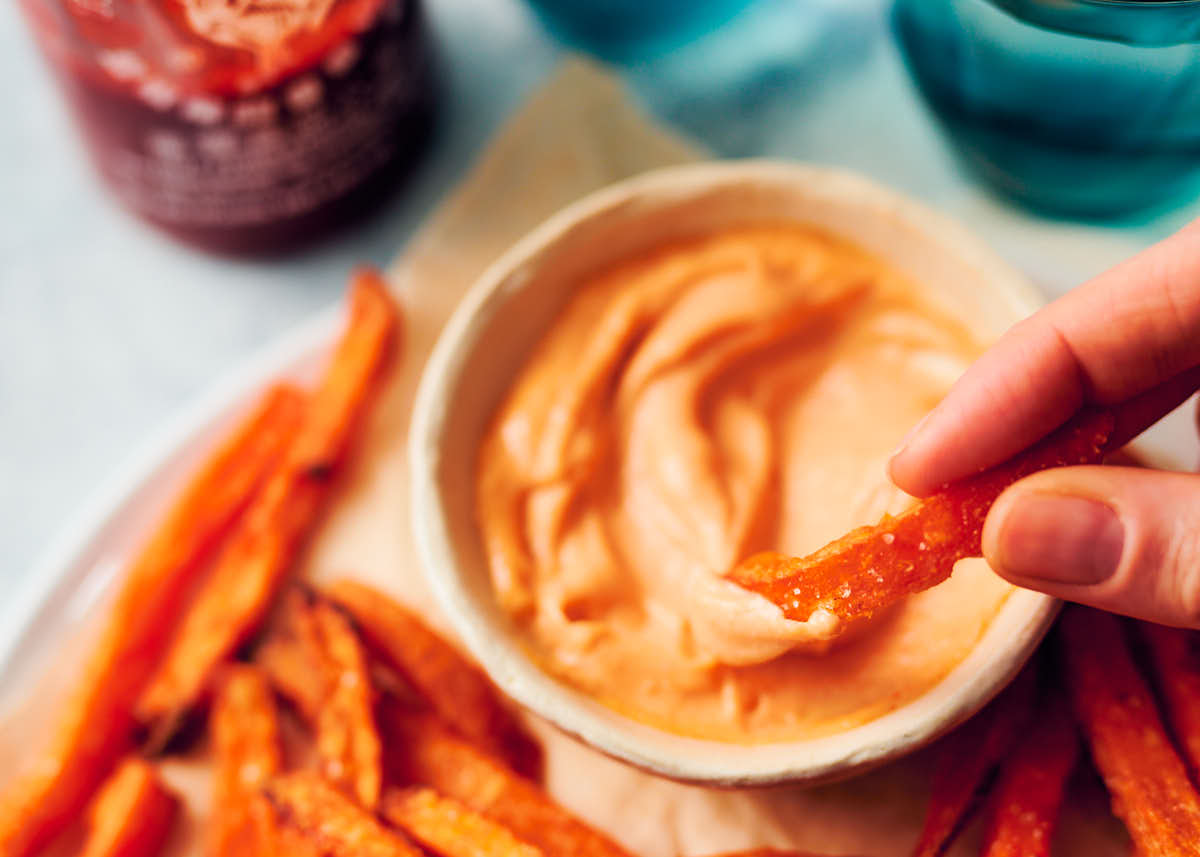 A sweet potato fry being dipped into a bowl of sriracha aioli.