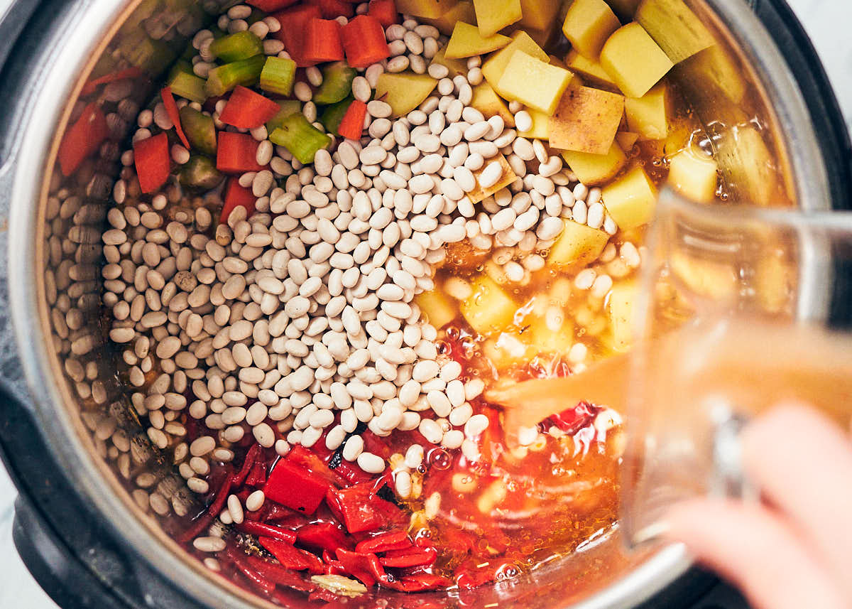 An Instant Pot with navy beans, roasted red peppers, carrots, celery, potatoes, and vegetable stock being poured in for Instant Pot Bean Soup.
