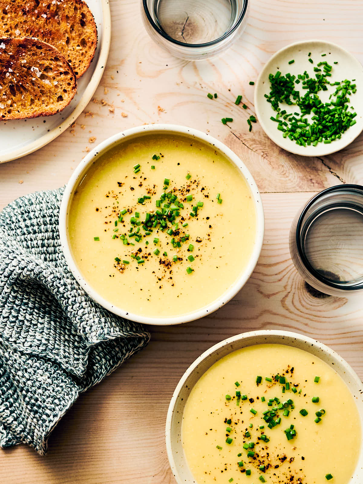 Two bowls of vegan potato leek soup on a table with bread and chives for serving.
