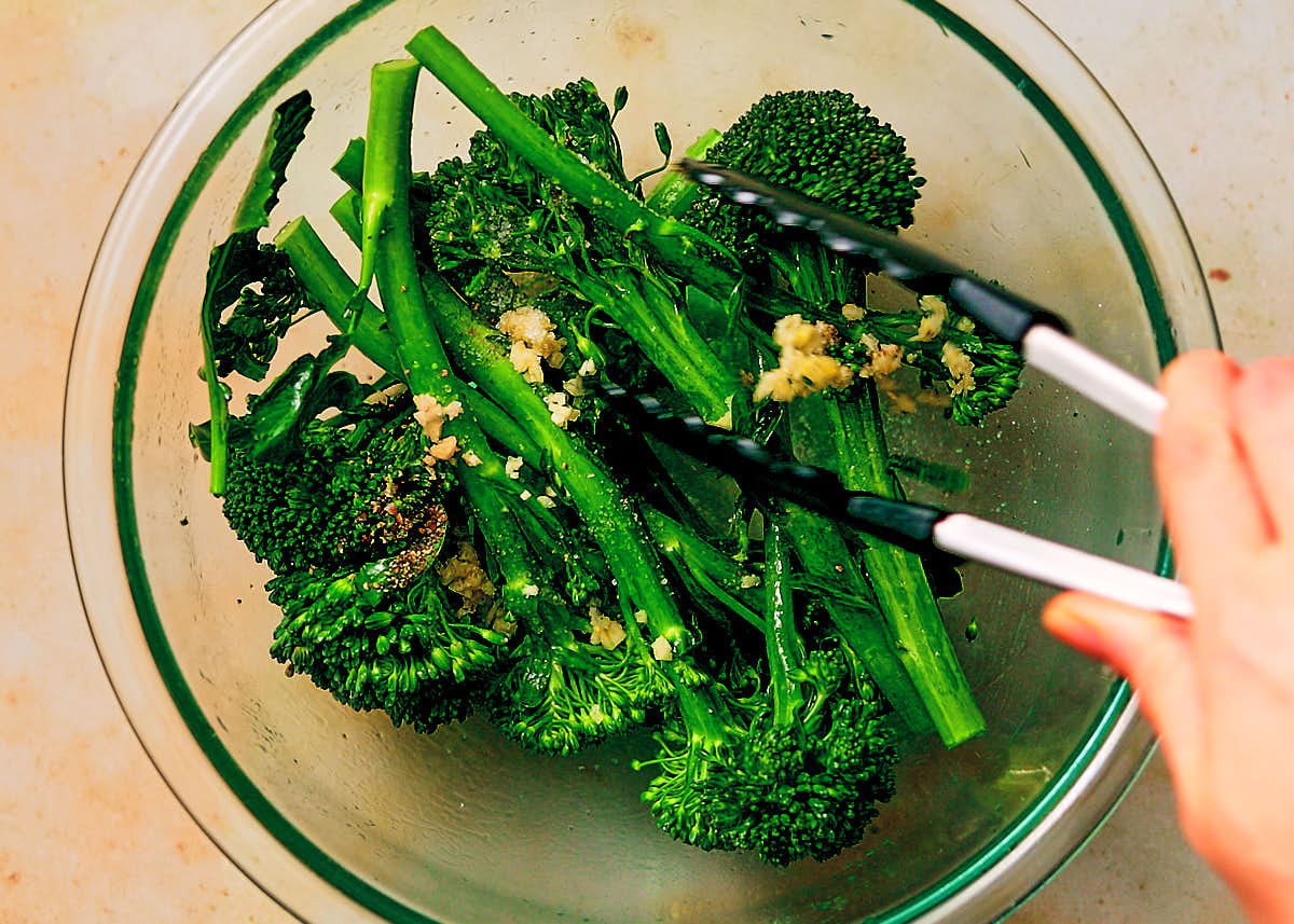 Tossing broccolini with oil, garlic, salt and pepper in a bowl with tongs before cooking.