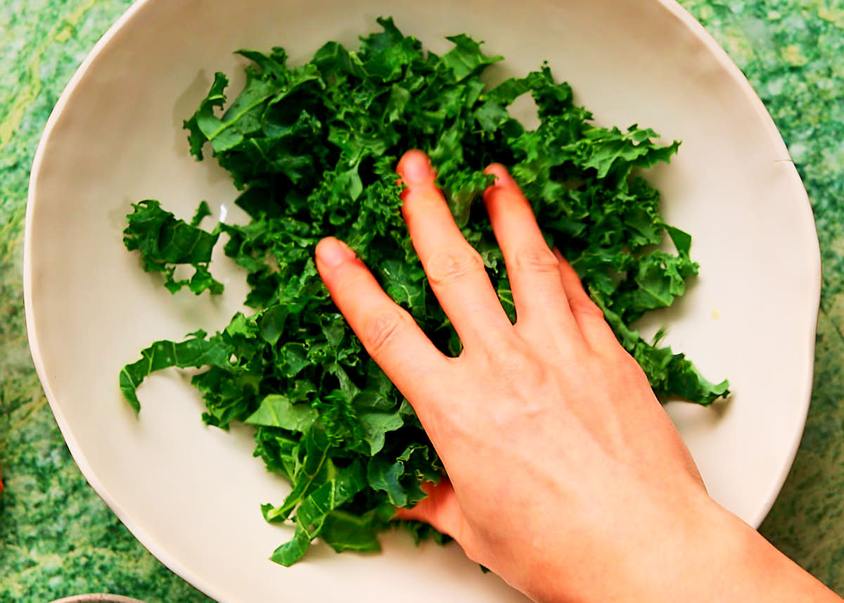 Hands massaging kale with olive oil and lemon juice in a bowl to soften.