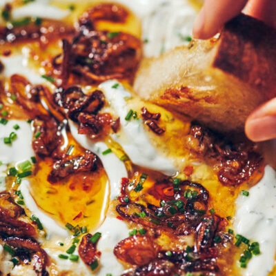 A piece of grilled bread being dipped into Greek yogurt onion dip with chili garlic oil and fried shallots
