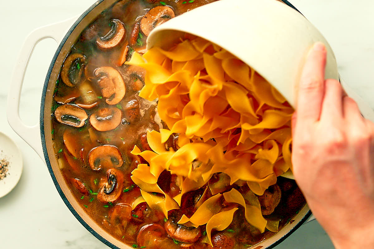 Adding noodles to simmering broth to make One Pot Mushroom Stroganoff.