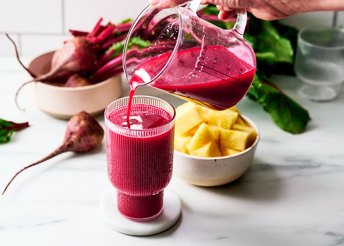 Beet Pineapple Cucumber Juice being poured into a glass with fresh beet and pineapple in bowls.
