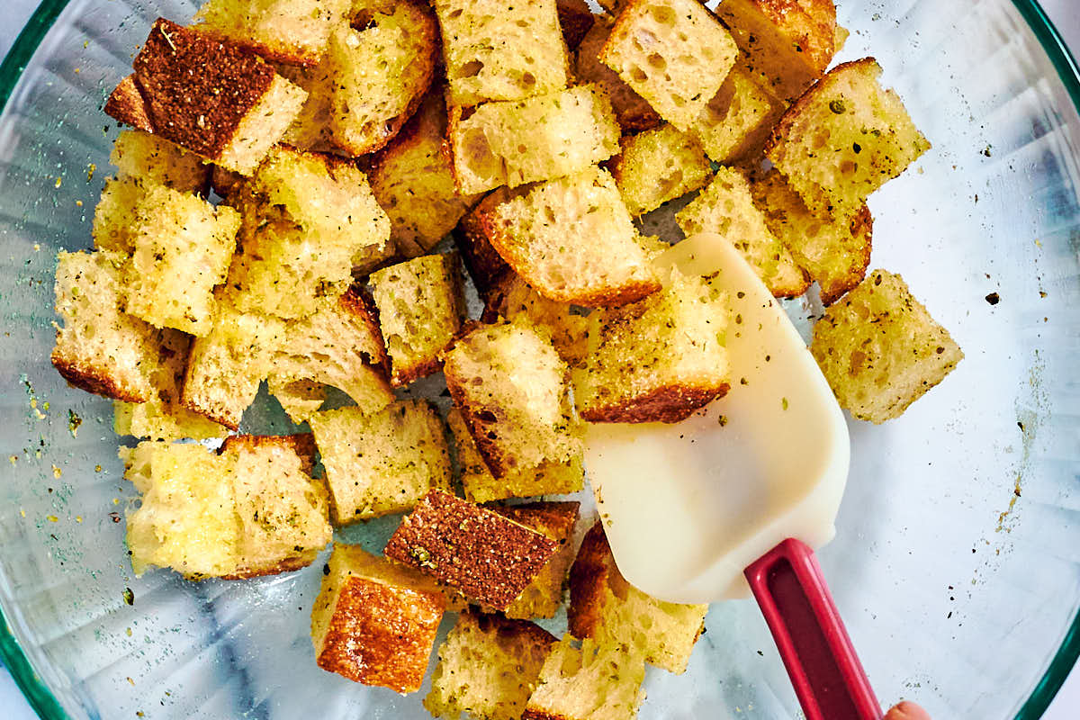 Seasoned bread cubes for croutons in a bowl with stirring spoon.