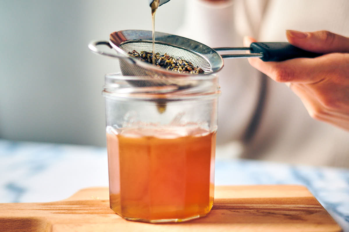 Pouring lavender and honey through a strainer over a glass jar.