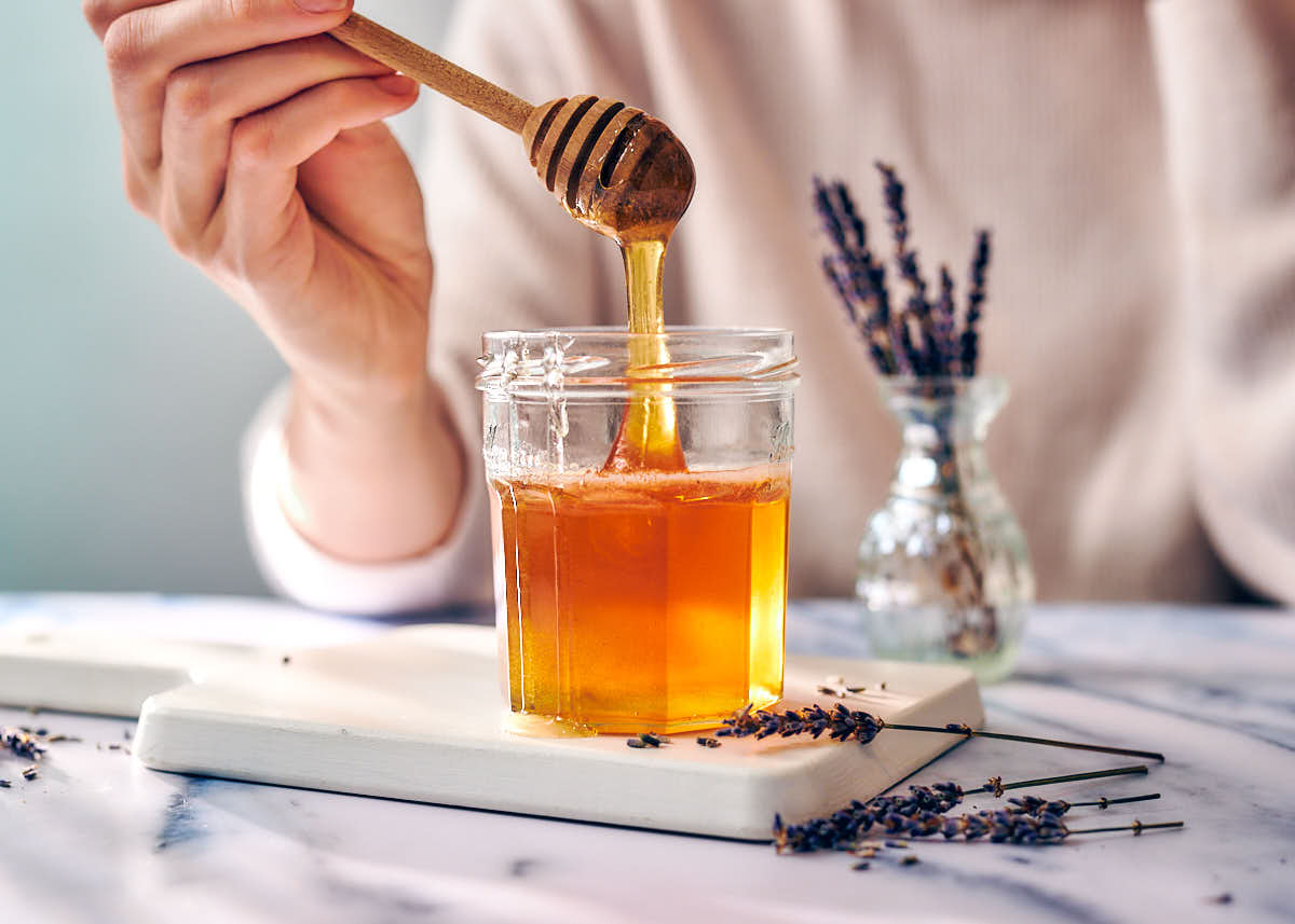 A woman spooning lavender honey out of a jar with a honey stick.