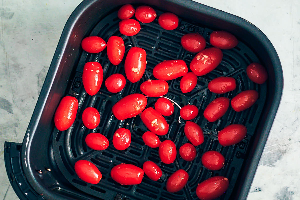Air fryer tomatoes tossed with olive oil and garlic in an air fryer basket before baking.