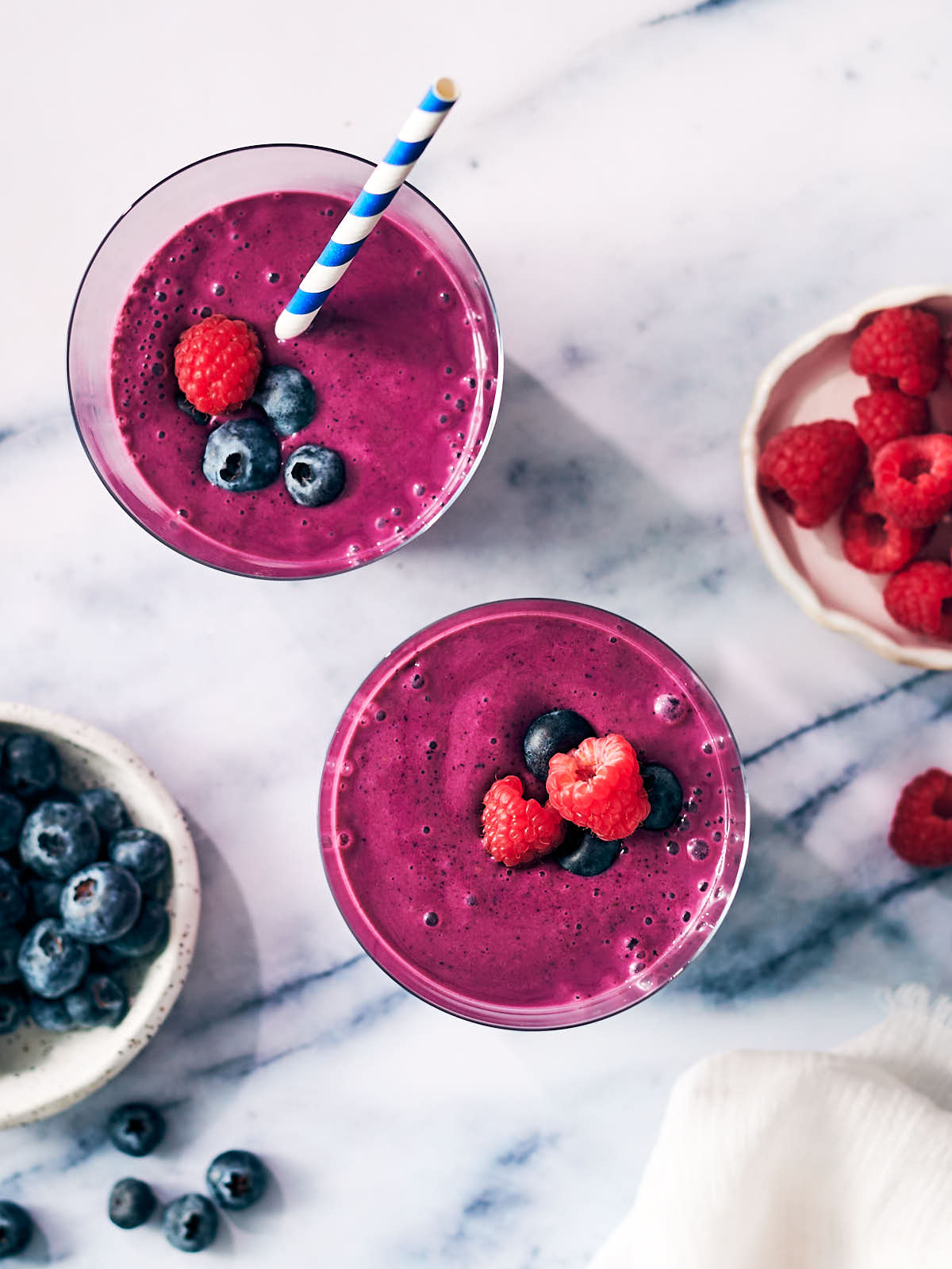 Two blueberry raspberry smoothies topped with fresh berries in a glass on a marble counter.