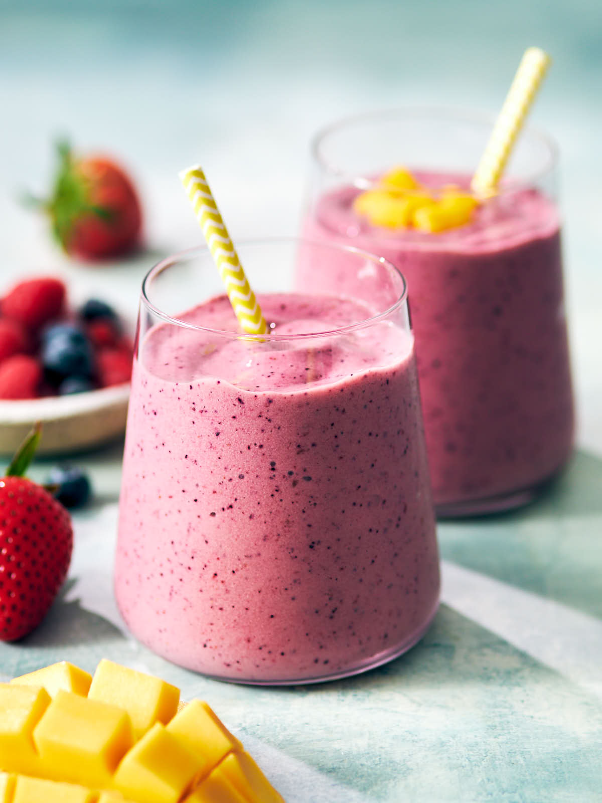Mango berry smoothies in glasses with straws surrounded by fresh fruit.